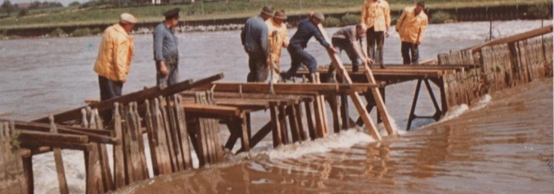 Das Foto zeigt einen Arbeitseinsatz am Nadelwehr der alten Kleinostheimer Schleuse (erbaut 1915 – 1917) im Jahr 1964. Ein Nadelwehr besteht aus einer Reihe von in den Fluss gesetzten Holzplanken (Nadeln). Die Durchflussmenge wird durch Wegnehmen oder Hinzufügen von Nadeln reguliert. Die Nadeln mussten regelmäßig herausgenommen und gereinigt werden. Durch den Neubau der Kleinostheimer Schleuse 1966 – 1971 wurde das Nadelwehr überflüssig.