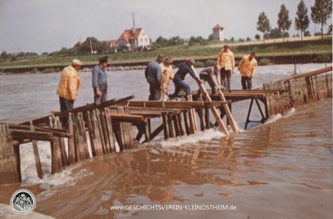 Das Foto zeigt einen Arbeitseinsatz am Nadelwehr der alten Kleinostheimer Schleuse (erbaut 1915 – 1917) im Jahr 1964. Ein Nadelwehr besteht aus einer Reihe von in den Fluss gesetzten Holzplanken (Nadeln). Die Durchflussmenge wird durch Wegnehmen oder Hinzufügen von Nadeln reguliert. Die Nadeln mussten regelmäßig herausgenommen und gereinigt werden. Durch den Neubau der Kleinostheimer Schleuse 1966 – 1971 wurde das Nadelwehr überflüssig.