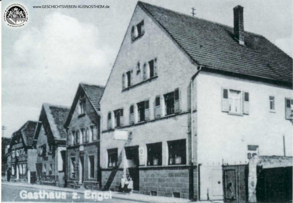 Das Foto zeigt das Gasthaus zum Engel im Jahre 1939. Erster Engelwirt war seit 1831 Peter Christ. Nach ihm folgten 1867 Johann Leitz, 1898 Johann Nikolaus Leitz und 1929 Cornelius Leitz. Der Saal hinter dem „Engel“ war erst Tanzsaal, dann in den 50er und frühen 60er Jahren das einzige Kino Kleinostheims (Gloria Lichtspiele). In den letzten Jahren hat sich der Name der Gaststätte wiederholt geändert.