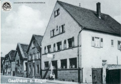 Das Foto zeigt das Gasthaus zum Engel im Jahre 1939. Erster Engelwirt war seit 1831 Peter Christ. Nach ihm folgten 1867 Johann Leitz, 1898 Johann Nikolaus Leitz und 1929 Cornelius Leitz. Der Saal hinter dem „Engel“ war erst Tanzsaal, dann in den 50er und frühen 60er Jahren das einzige Kino Kleinostheims (Gloria Lichtspiele). In den letzten Jahren hat sich der Name der Gaststätte wiederholt geändert.